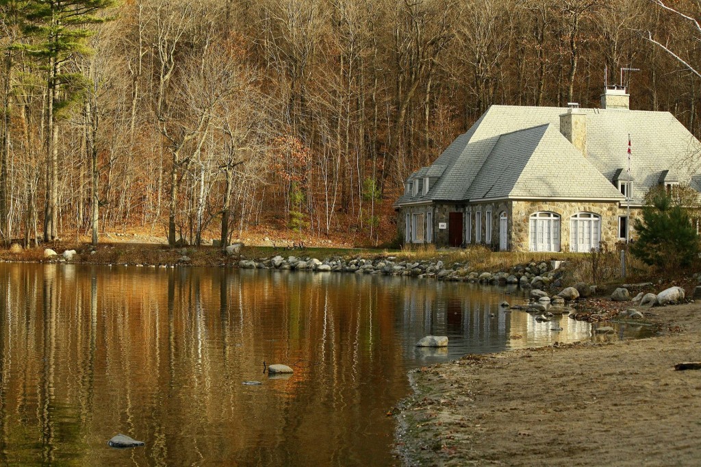 Image of a chalet next to a lake. The trees in the background are bare and leaves cover the ground; it appears to be late fall.